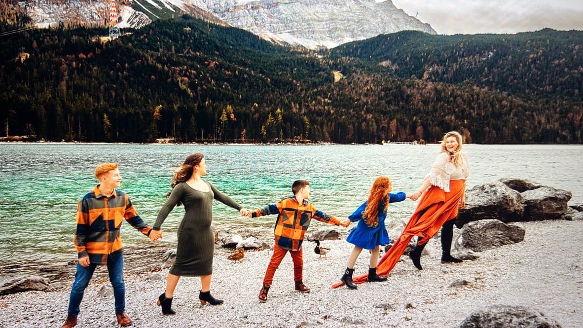A family of five holding hands in a line along a lake shore, mountains behind them.