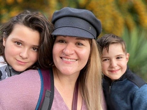 A woman in a cap between two children, all three smiling, autumn leaves behind.