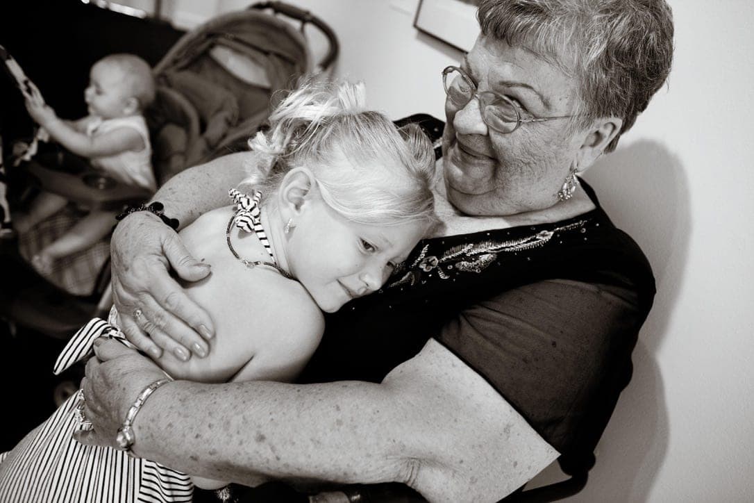 An older woman holding two small children close, her eyes shut. Black and white.