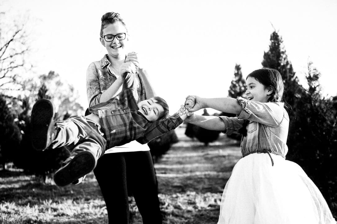 Two adults swinging a child between them by the arms, a girl in a white dress reaching for him. Black and white, outdoors.