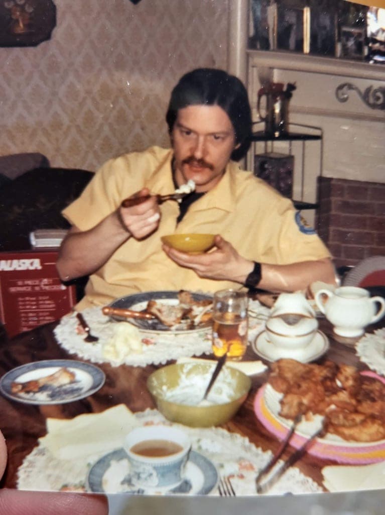 A man in a yellow work shirt eating at a laid table — good china, a patterned cloth, a bowl held in one hand.