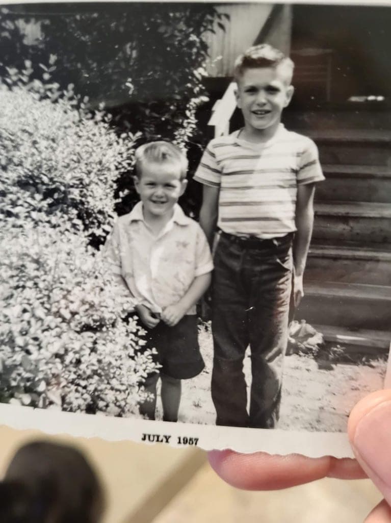 Two boys in striped shirts beside a flowering bush on a summer step. JULY 1957 is printed on the border, and a hand holds the corner of the print.